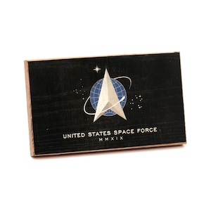 May include: A rectangular black wooden plaque featuring the United States Space Force emblem. The design includes a silver delta, a blue globe with grid lines, and white orbital rings. The text reads UNITED STATES SPACE FORCE and MMXIX.