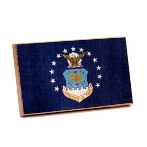 May include: A rectangular wooden plaque featuring the United States Air Force emblem. The plaque has a dark blue background with the emblem in the center, surrounded by white stars. The words "United States Air Force" are visible.