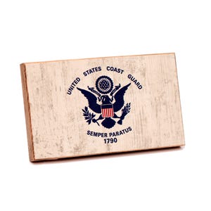 May include: A rectangular wooden plaque with a distressed off-white finish featuring the United States Coast Guard emblem in dark blue, red, and white. The text reads UNITED STATES COAST GUARD, SEMPER PARATUS, and the year 1790 in black lettering.