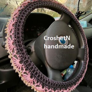 May include: Crochet steering wheel cover in pink and black with a delicate lace pattern. The cover is handmade and says "CroshetN handmade".