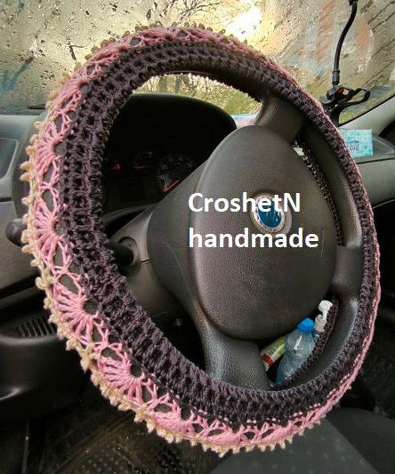 May include: Crochet steering wheel cover in pink and black with a delicate lace pattern. The cover is handmade and says "CroshetN handmade".