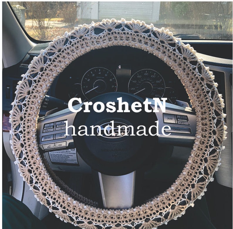 May include: Crochet steering wheel cover in beige and white with a delicate lace pattern. The cover is handmade and features the text "CroshetN handmade".