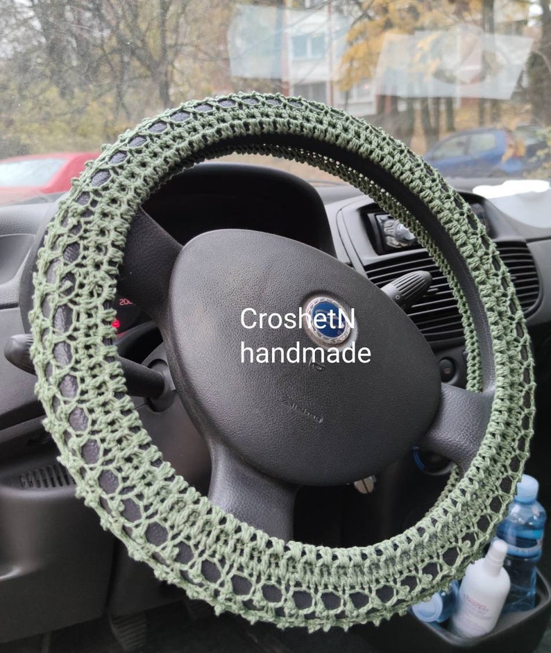 May include: Green crocheted steering wheel cover. The cover is made with a delicate open stitch pattern. The text "CroshetN handmade" is visible on the steering wheel.