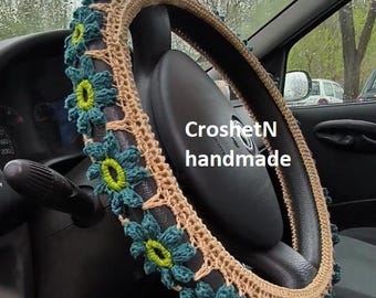 May include: Crochet steering wheel cover with beige and teal blue flowers. The cover is handmade and has the text "CroshetN handmade" on it.