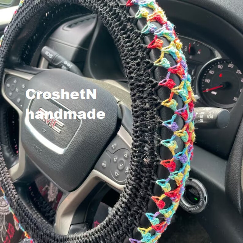 May include: A black crochet steering wheel cover with a colorful rainbow design. The cover is handmade and has the text "CroshetN handmade" on it.