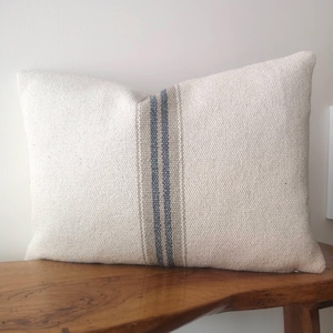 May include: Rectangular off-white pillow with a vertical stripe in the center. The stripe features a combination of blue and tan colors. The pillow is placed on a wooden bench, showcasing a rustic aesthetic.