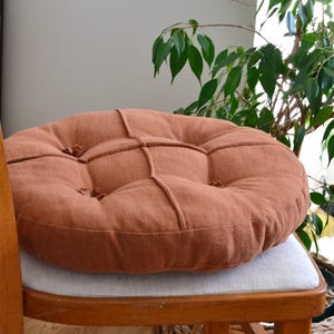 May include: A round, rust-colored cushion with a tufted design sits on a wooden chair. The cushion is made of a textured fabric and has a stitched pattern. The chair is made of wood and has a light-colored seat.