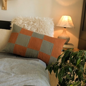 Op de afbeelding: Een rechthoekig kussen met een patchwork ontwerp in oranje en grijstinten. Het kussen ligt op een bed met grijs beddengoed, aangevuld met een fluffy wit kussen. Een lamp en een plant zijn zichtbaar op de achtergrond.