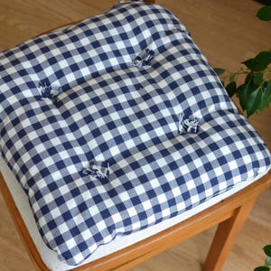 May include: A square cushion with a blue and white gingham pattern, button tufting, and a light-colored fabric base. The cushion is placed on a wooden chair with a curved back and legs.