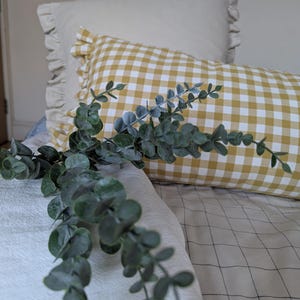 May include: A yellow and white gingham pillow with frills sits on a bed with a light grey and black checked bedspread. A sprig of green eucalyptus is placed on the bed.