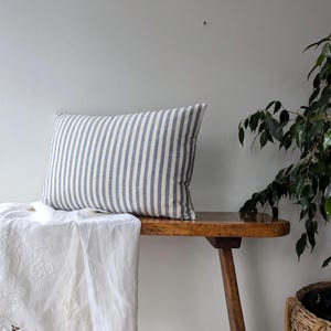 Op de afbeelding: Een kussen met blauwe en witte strepen ligt op een houten bank. Het kussen heeft een rechthoekige vorm. Een wit geborduurde doek is over de bank gedrapeerd. Een groene plant staat op de achtergrond.