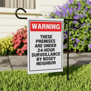 Puede incluir: Un cartel de advertencia blanco con texto rojo que dice "WARNING" y texto negro que dice "THESE PREMISES ARE UNDER 24-HOUR SURVEILLANCE BY NOSEY NEIGHBOR". El cartel está sujeto a una estaca de jardín de metal negro.