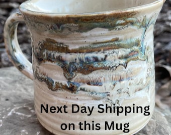 May include: A handmade stoneware mug with a unique, textured design. The mug features a cream-colored base with streaks of brown and blue. The handle is curved, and the rim is slightly flared.