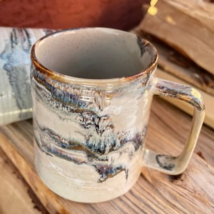 Peut inclure: Une tasse en céramique avec un glaçage blanc et des marbrures brunes, bleues et blanches. La tasse a une grande anse et une large ouverture.