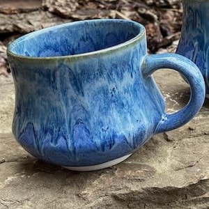 Peut inclure: Deux tasses en céramique présentant un vernis bleu brillant aux tons multiples avec un motif de coulures distinctif. Chaque tasse possède un corps arrondi, un bord large et une anse incurvée. Le vernis passe du bleu roi au bleu ciel.