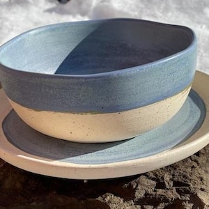 May include: A handmade ceramic bowl and saucer set. The bowl is a light blue color on the top half and a cream color on the bottom half. The saucer is cream with a light blue center. The set is sitting on a rock.