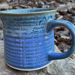 May include: A handmade ceramic mug with a blue and teal glaze. The mug has a rounded handle and a textured surface with horizontal lines. The mug is sitting on a grey rock.