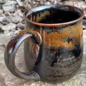 May include: A ceramic mug featuring a rounded body and a curved handle. The exterior surface displays a reactive glaze with a deep black base colour transitioning into a warm, earthy amber and brown drip pattern near the rim and handle area.