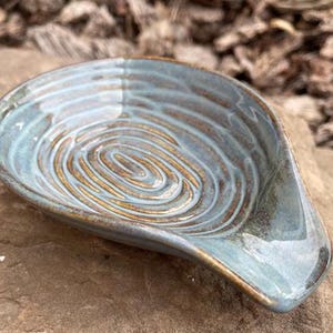 Peut inclure: Un repose-cuillère en céramique fait à la main avec un glaçage bleu-vert et un motif en spirale au centre. Le repose-cuillère a une forme incurvée et repose sur une surface en pierre. Le repose-cuillère mesure environ 15 cm de long.