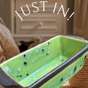 May include: Green ceramic loaf pan with black speckled glaze and black handles.  "Just In!" is written in white text above the pan.