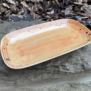 May include: A rectangular ceramic tray with rounded corners, featuring a glazed finish in shades of warm orange and tan. The surface displays visible brushstroke textures and scattered dark blue and black speckles along the edges and rim.