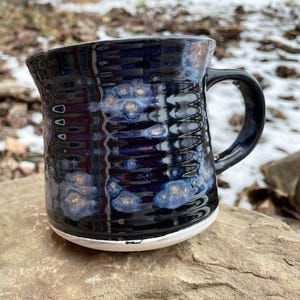 May include: A handmade ceramic mug with a black and blue glaze. The mug has a textured surface and a white base. The interior is a dark blue color. The mug is sitting on a rock.