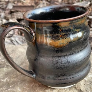May include: A ceramic mug with a rounded, wavy silhouette and a glossy finish. The exterior features a blend of deep black and metallic bronze tones, with a prominent curved handle on the side. The rim is smooth and circular, showing a dark glaze.