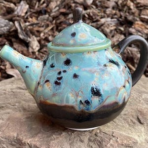 May include: A ceramic teapot featuring a glossy glaze with a blend of turquoise, light blue, and dark brown colours. The teapot has a rounded body, a curved spout, a dark brown handle, and a matching lid with a small knob on top.