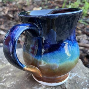 May include: A ceramic mug featuring a glossy glaze with a colour gradient transitioning from deep navy blue at the rim to a vibrant turquoise in the middle, and a warm earthy brown at the base. The mug has a curved handle and a white circular base.