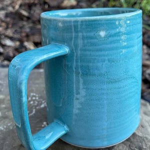 Peut inclure: Une tasse en céramique cylindrique avec un émail turquoise brillant. La surface présente de subtiles stries horizontales et une anse plate et lisse fixée sur le côté. L'émail montre des variations d'intensité, allant du bleu-vert clair au foncé.