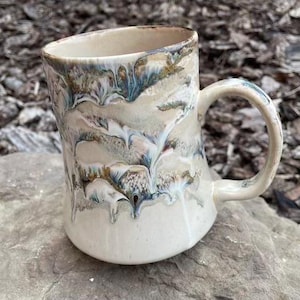 Peut inclure: Mug en céramique fait à la main avec un glaçage unique. La base est crème avec des motifs fluides bleus, bruns et blancs. La poignée est incurvée et le bord est irrégulier. Idéal pour le café ou le thé.