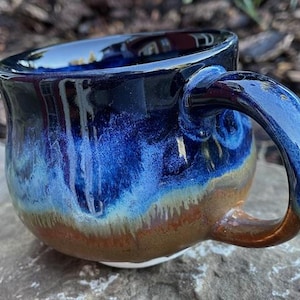 Peut inclure: Une tasse en céramique avec un corps arrondi et une anse incurvée. L'émail présente un bleu marine profond sur le dessus qui passe à un bleu ciel vibrant, se fondant dans un brun terreux chaud à la base avec une finition brillante.