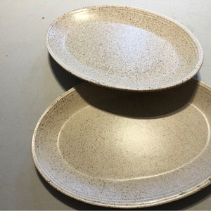 May include: Two oval ceramic serving platters. The plates have a speckled, off-white glaze with dark brown speckles. The rims have a slightly raised edge and a darker brown trim. The plates are stacked on a grey surface.
