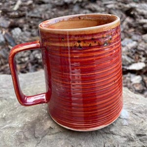 May include: A red ceramic mug with a striped pattern. The mug has a wide handle and a rounded shape.