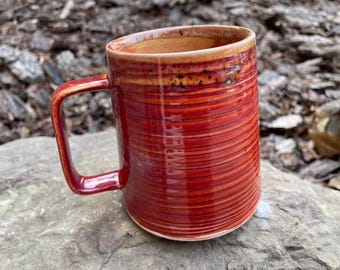 May include: A red ceramic mug with a striped pattern. The mug has a wide handle and a rounded shape.
