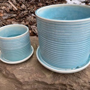 May include: Two light blue ceramic plant pots with matching saucers. The pots have a ribbed texture and a white rim. The larger pot is approximately 18 cm tall, and the smaller one is about 10 cm tall. They are sitting on a large rock.
