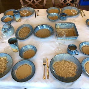 May include: A table setting with blue and brown ceramic plates, bowls, mugs, and a casserole dish. The dishes have a speckled glaze with a light brown color and a blue rim.