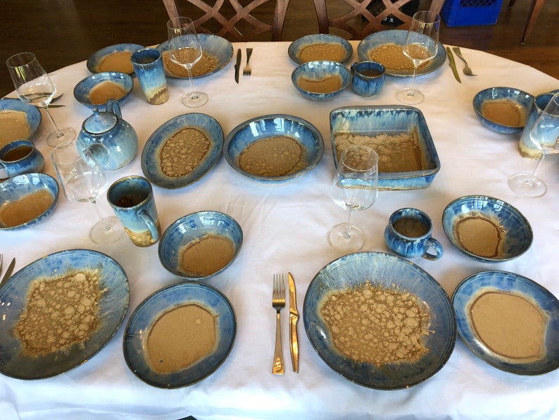 May include: A table setting with blue and brown ceramic plates, bowls, mugs, and a casserole dish. The dishes have a speckled glaze with a light brown color and a blue rim.