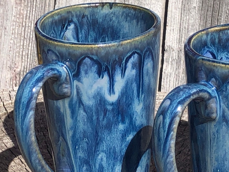 May include: Two blue and white ceramic mugs with a drip glaze finish. The mugs have a handle and are tall and slender.