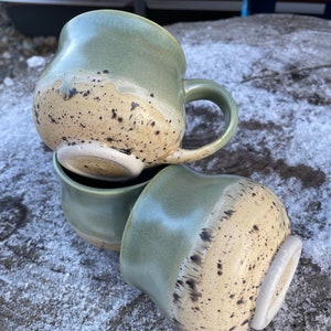 May include: Three ceramic mugs stacked on top of each other. The mugs are green with a speckled glaze and a light brown band around the middle.