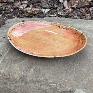 May include: An oval ceramic plate with a glossy finish, featuring a warm orange and tan centre that transitions to a darker, speckled brown rim. The plate has a smooth, slightly concave surface and a distinct, uneven edge texture.