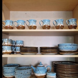 May include: A set of blue and brown speckled ceramic mugs, bowls, plates, and a teapot. The dishes are arranged on a wooden shelf in a kitchen cabinet.
