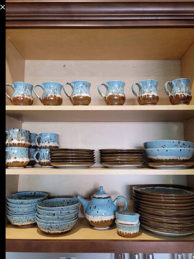 May include: A set of blue and brown speckled ceramic mugs, bowls, plates, and a teapot. The dishes are arranged on a wooden shelf in a kitchen cabinet.