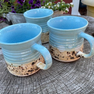 May include: Three blue ceramic mugs with a speckled brown and cream glaze. The mugs have a rounded shape and a handle on the side.