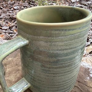May include: A handmade ceramic mug with a large handle. The mug has a green and grey marbled glaze. The interior and rim are a lighter green. The mug is sitting on a stone surface.