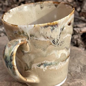 Peut inclure: Une tasse en céramique faite à la main avec un design unique et organique. La tasse présente une base beige avec des motifs fluides de blanc, de marron et de vert. La poignée est également ornée de couleurs similaires. La tasse a un bord légèrement incliné et une base blanche.