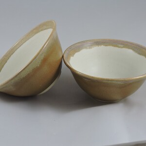 May include: Two ceramic bowls with a light brown glaze and a white interior. The bowls have a subtle, uneven glaze pattern.
