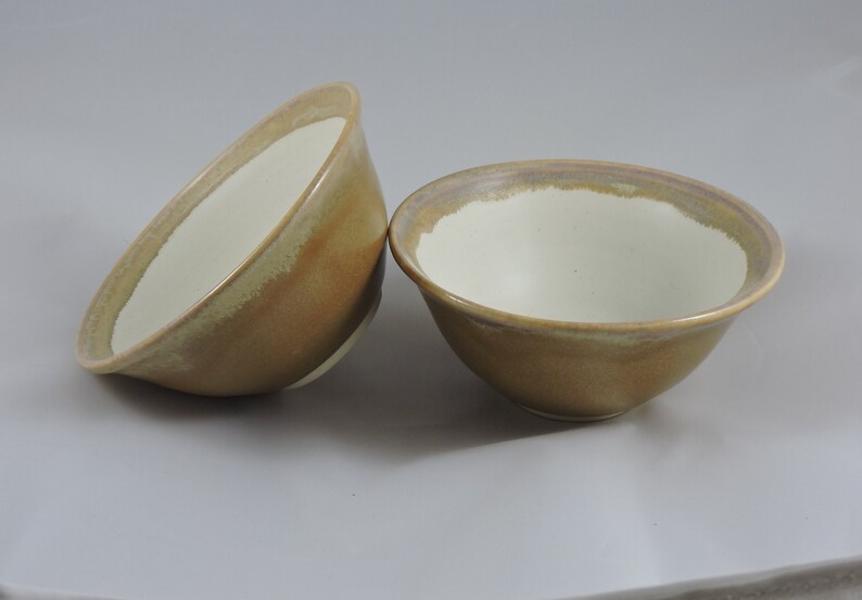 May include: Two ceramic bowls with a light brown glaze and a white interior. The bowls have a subtle, uneven glaze pattern.