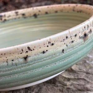 May include: A ceramic bowl with a light green glazed interior and exterior featuring horizontal ridges. The rim is finished in a contrasting cream glaze with dark brown speckles that bleed downward, creating a textured appearance along the edge.