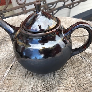 May include: A dark brown ceramic teapot with a glossy finish, featuring a lid and a curved handle. The teapot has a rounded body and a spout. The surface reflects light, highlighting the smooth texture and rich colour. A small knob sits atop the lid.